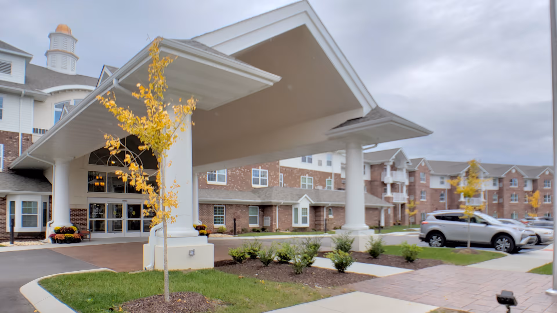 Entrance of Ashwood Square Retirement Community showing a covered drop-off area supported by large white columns, with a brick building in the background and a parking area with cars to the right. Small trees with yellow leaves and landscaped bushes are visible around the entrance.