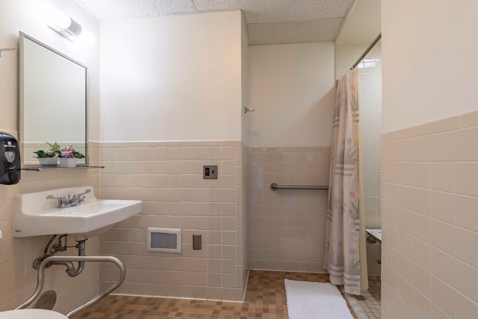 A clean bathroom with beige tiled walls and a checkered floor. The room features a white sink with a mirror above it, a soap dispenser on the wall, a small shelf with potted plants, a shower area with a floral-patterned shower curtain, and a white bath mat on the floor. There is a grab bar installed near the shower for accessibility.