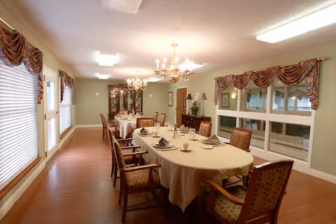 A well-lit dining room with a long oval table covered with a white tablecloth, set with plates, cups, and napkins. The room has wooden floors, large windows with decorative curtains, chandeliers hanging from the ceiling, and a china cabinet in the background.