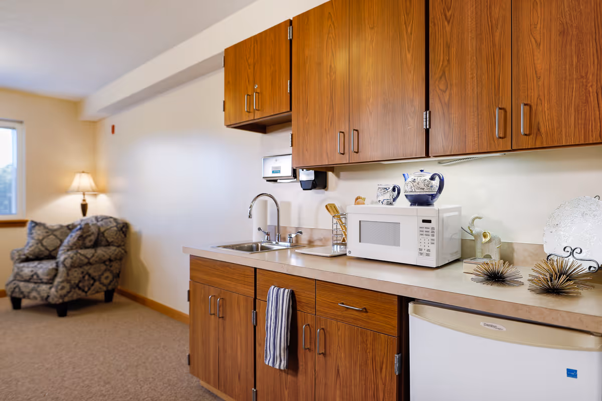 Small kitchenette with wooden cabinets, a sink, microwave and mini-fridge next to a sitting area with an upholstered armchair and lamp.
