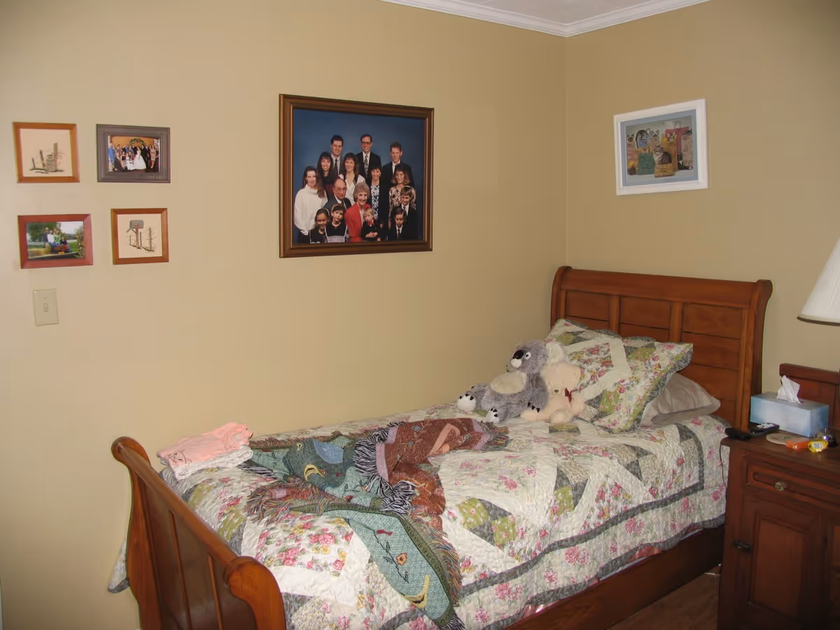 A cozy bedroom with a wooden bed frame and a floral quilted bedspread. There are two stuffed animals on the bed, a gray and white one and a beige teddy bear. On the wall above the bed are several framed pictures, including a large family portrait and smaller photos. A wooden nightstand next to the bed holds a lamp, a tissue box, and some small items.