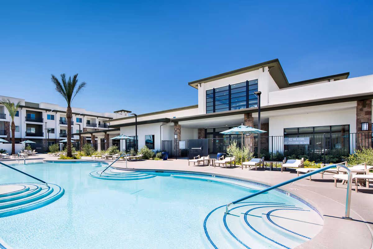 Outdoor swimming pool area at Revel Nevada facility with clear blue water, curved steps leading into the pool, lounge chairs with umbrellas, palm trees, and a modern building in the background under a clear blue sky.