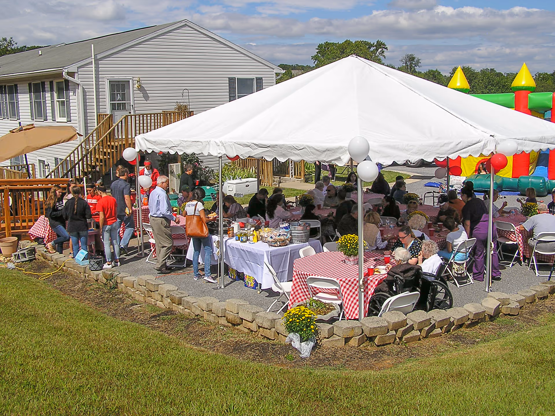 Outdoor gathering at Country Acres Assisted Living with people seated under a white canopy tent at tables covered with red and white checkered tablecloths. There are balloons tied to the tent poles, a buffet table with food and drinks, and a colorful inflatable bounce house in the background. The event is set on a paved area next to a building with a grassy lawn in the foreground.