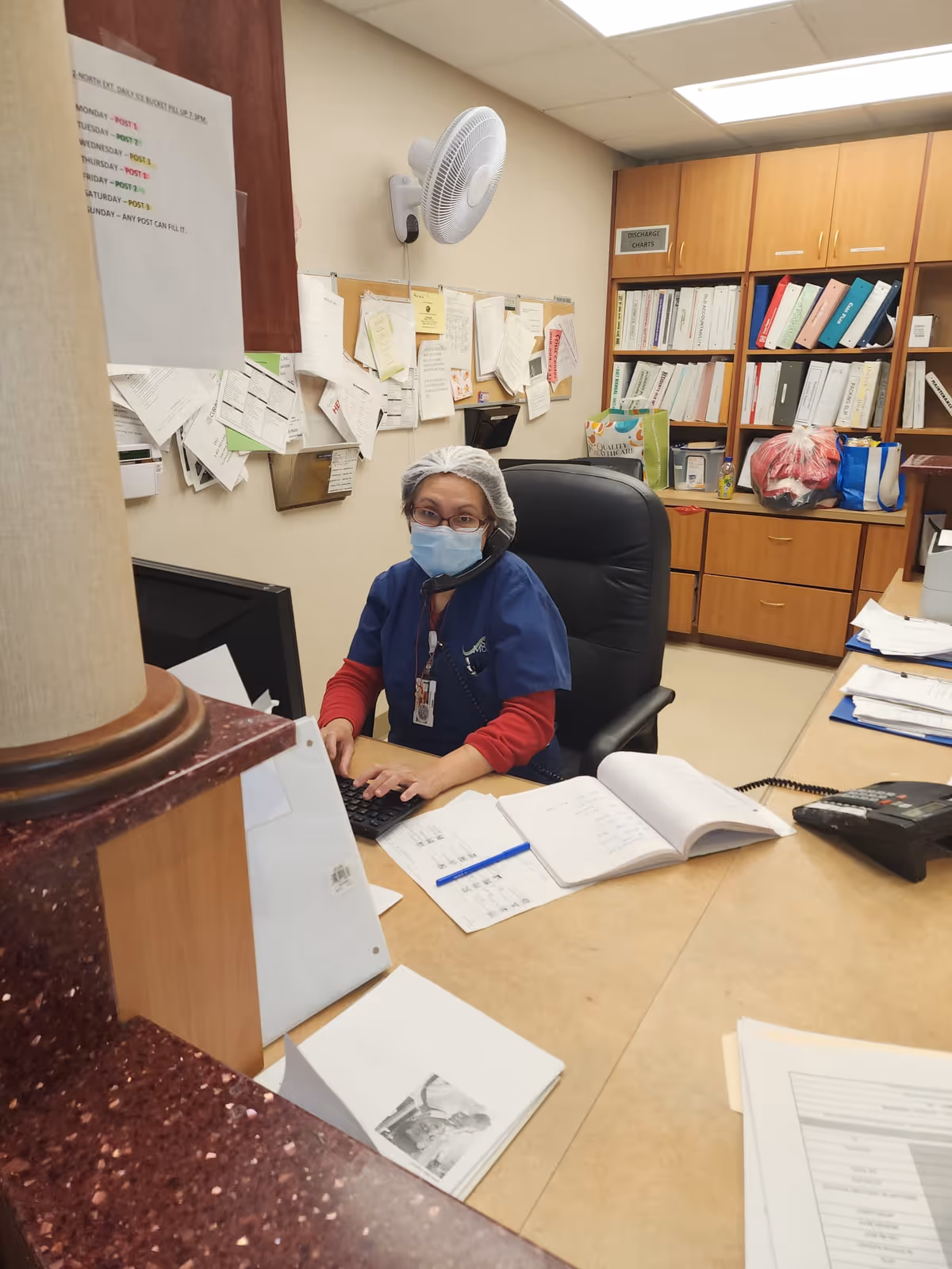 A healthcare worker wearing a hairnet, face mask, and blue scrubs is sitting at a desk in an office area. The worker is using a keyboard and talking on a phone headset. The desk is cluttered with papers, notebooks, and a telephone. Behind the worker, there are shelves filled with binders and various office supplies, and a bulletin board with pinned papers on the wall.