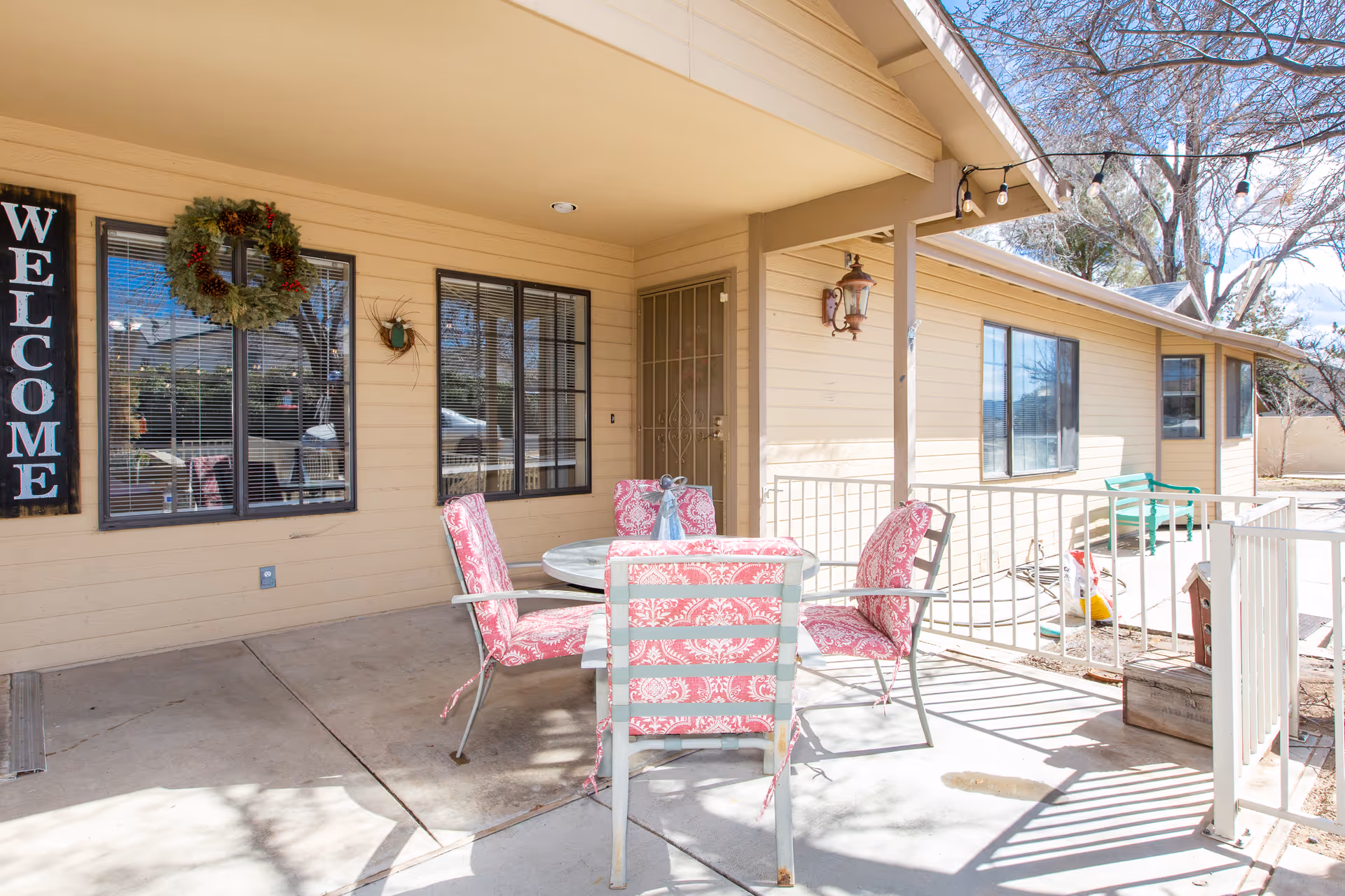 Outdoor patio area of Prescott Valley Assisted Living with a round table and four chairs with pink patterned cushions. The building exterior is beige with windows and a door. A large vertical 'WELCOME' sign and a wreath are mounted on the wall. There are string lights hanging above and a tree with bare branches nearby.