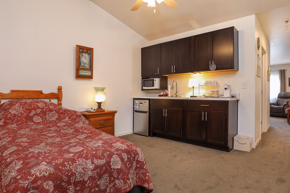 A cozy assisted living room featuring a wooden bed with a red floral bedspread, a wooden nightstand with a glowing lamp, and a kitchenette area with dark wood cabinets, a microwave, mini fridge, sink, and a small lamp. The room has beige walls and carpeted floor, with a hallway leading to another room with a recliner chair visible.