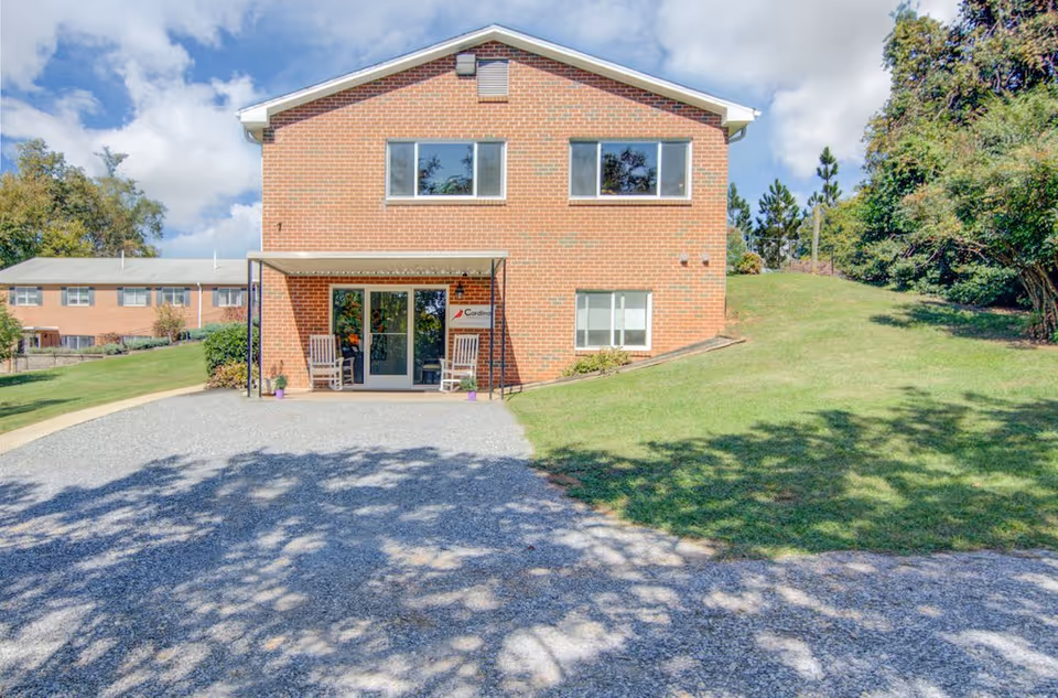 Front exterior view of a two-story brick building with a small covered entrance featuring two rocking chairs and potted plants. The building is surrounded by green grass, trees, and a gravel driveway under a partly cloudy sky.