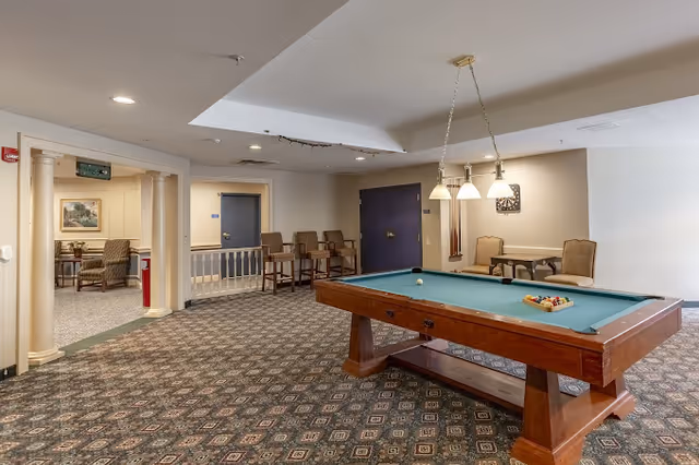 A communal recreation room with a pool table, hanging lights, patterned carpet and seating areas.
