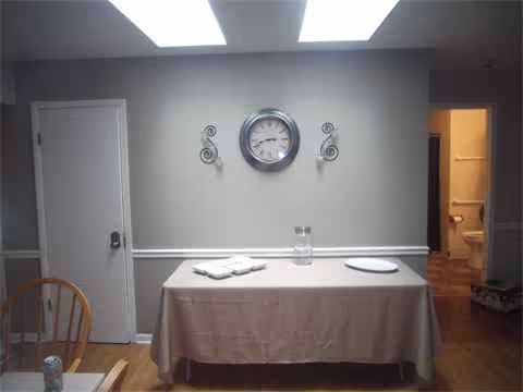 A long table with a tablecloth, napkins, and a clock on the wall in dining area.