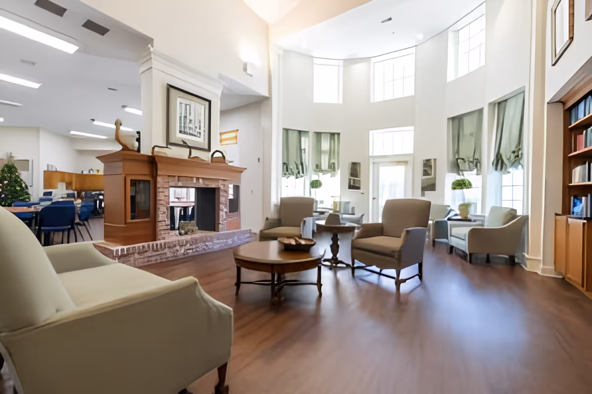 A bright and spacious living room area with high ceilings and large windows allowing natural light to fill the space. The room features comfortable armchairs and sofas arranged around wooden coffee tables. There is a fireplace with a wooden mantle and framed artwork above it. In the background, a dining area with tables and chairs is visible, along with a decorated Christmas tree. Shelves with books and decorative items are on the right side of the room.