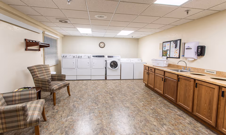 Communal laundry room with washers and dryers, a sink and cabinets along one wall, and two plaid armchairs.