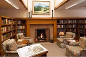 Cozy library room with a lit fireplace in the center, surrounded by wooden bookshelves filled with books. Four upholstered armchairs with floral patterns are arranged around the fireplace, with a small table holding an open book in the foreground. A framed landscape painting hangs above the fireplace.