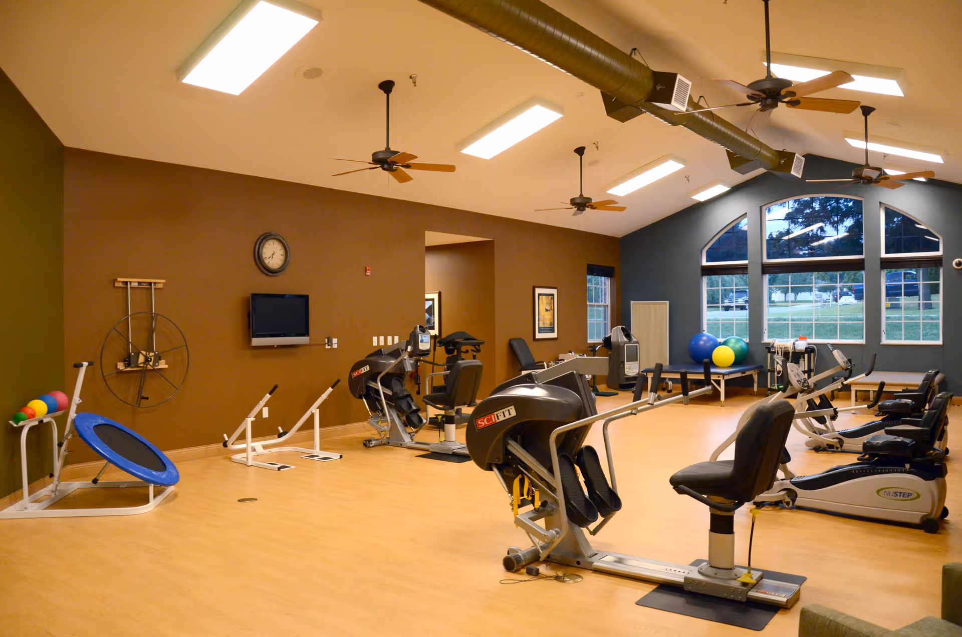 A spacious fitness room with exercise equipment including recumbent bikes, a small trampoline, exercise balls, and other workout machines. The room has a high ceiling with ceiling fans and large windows letting in natural light. The walls are painted brown and blue, and there is a clock and a wall-mounted TV on one wall.