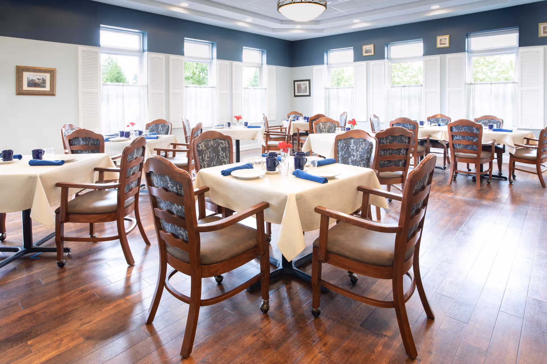 A bright dining room with multiple tables covered in beige tablecloths, each set with blue napkins, plates, and mugs. Wooden chairs with cushioned seats and backs surround the tables. Large windows with white shutters allow natural light to fill the room, and framed pictures hang on the walls.