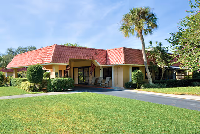 Exterior view of a single-story building with a red tiled roof, a covered entrance with chairs, surrounded by green grass, bushes, and a palm tree under a clear blue sky.