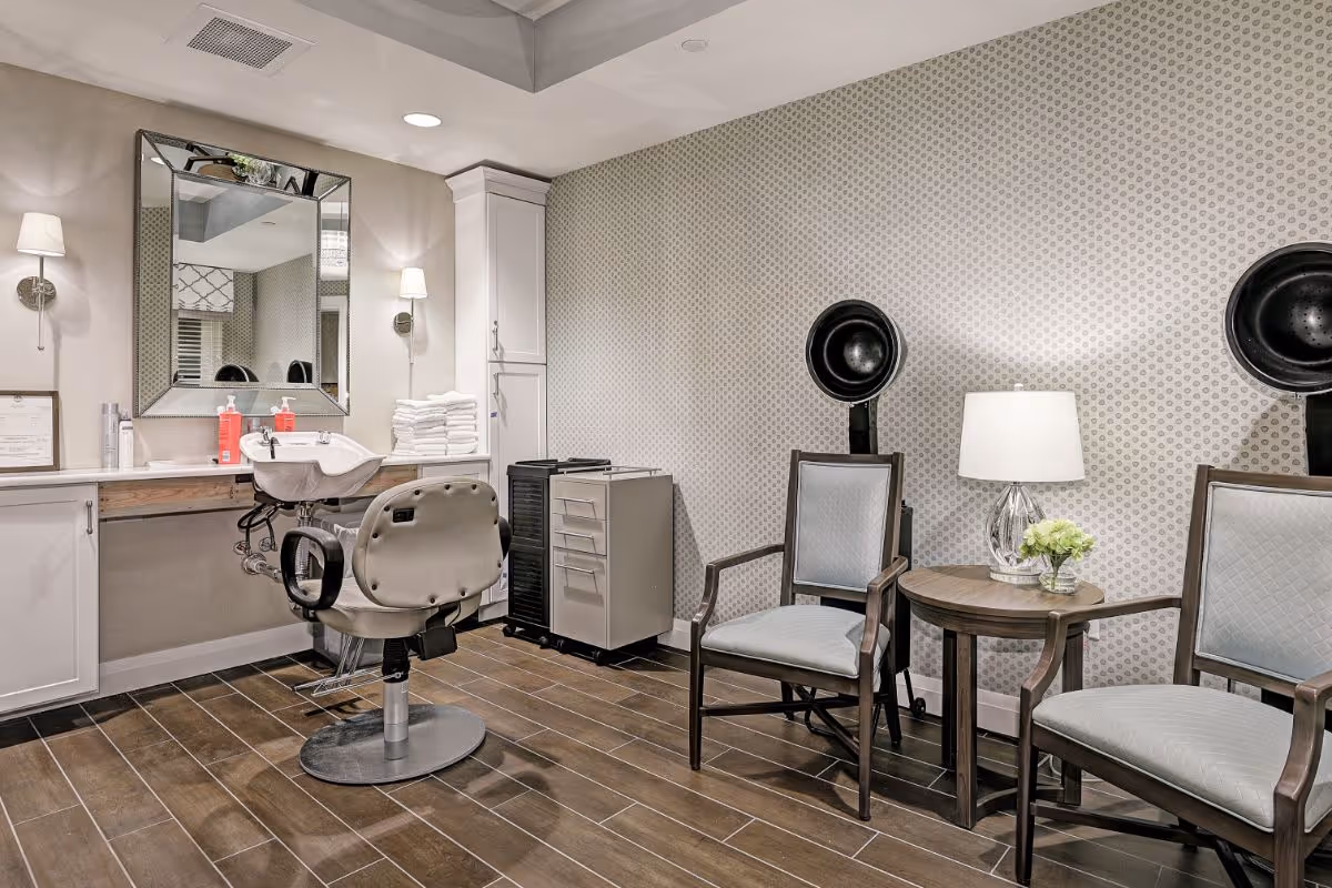 Salon-style interior with a styling chair and wash sink, two hooded hair dryers, and seating with a side table and lamp.