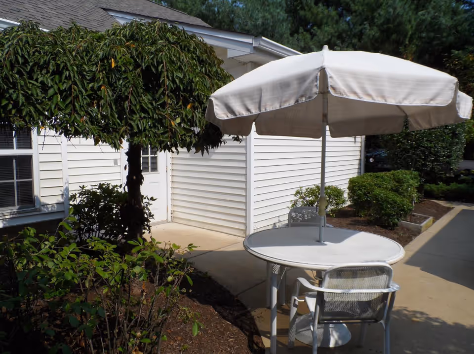 Outdoor patio area with a round white table and three metal chairs under a large white umbrella. The patio is adjacent to a white building with a door and window. There are green bushes and a tree with dense foliage nearby.