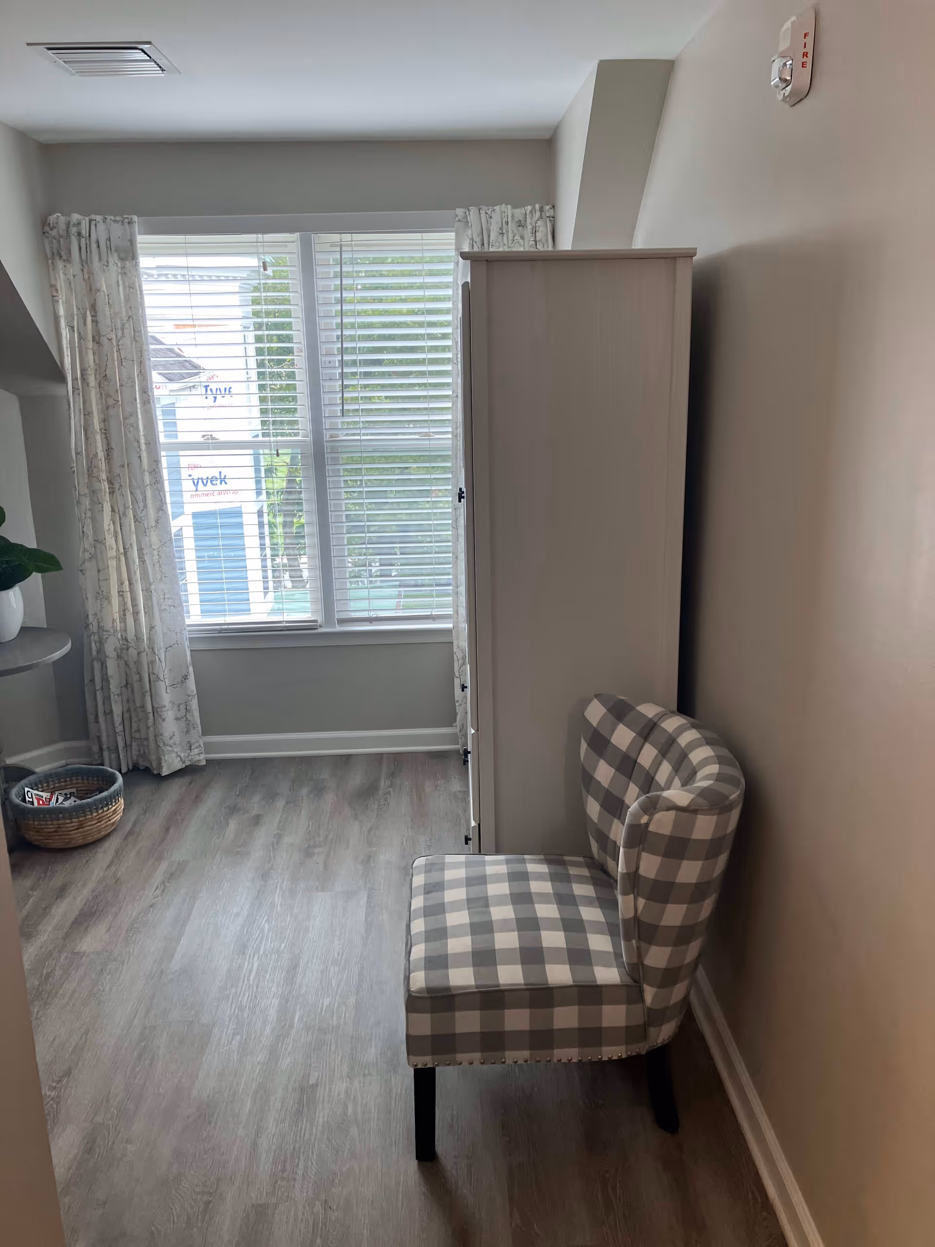 A small room with a checkered upholstered chair placed against a beige wall. There is a tall white cabinet next to the chair. A window with white blinds and patterned curtains lets in natural light. The floor is covered with light wood laminate. A small round table with a plant and a basket with magazines are visible in the corner near the window.