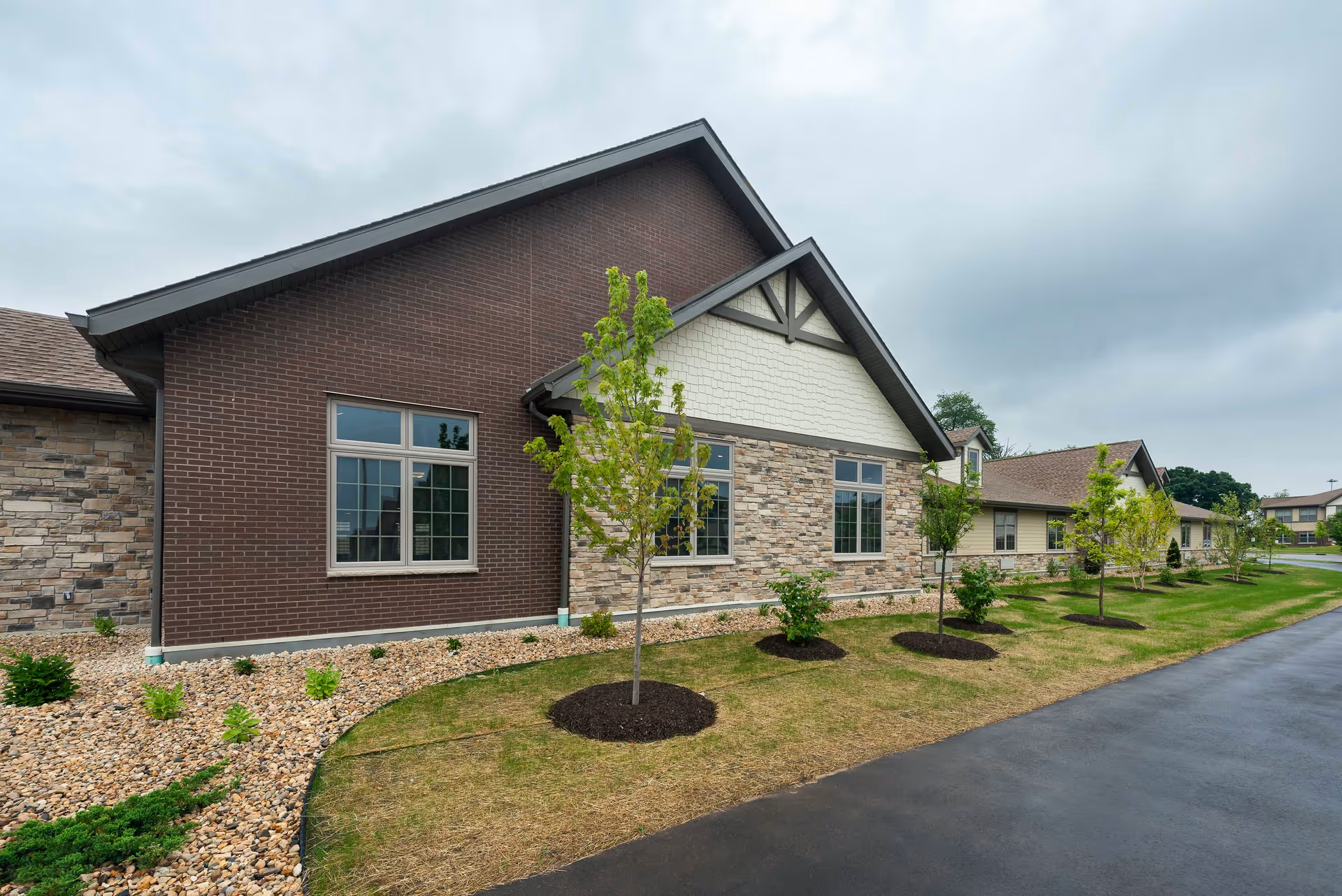 Exterior view of a senior living facility building with a combination of brick, stone, and siding on the walls. There are several young trees planted along a landscaped area with mulch and rocks, and a paved driveway or walkway runs alongside the building under a cloudy sky.
