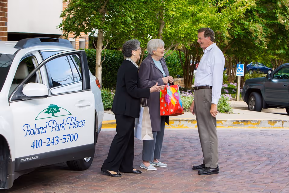 Three elderly people standing outside near a white car with the Roland Park Place logo and phone number on the door. Two women are holding shopping bags and talking with a man. Trees and parked cars are visible in the background.