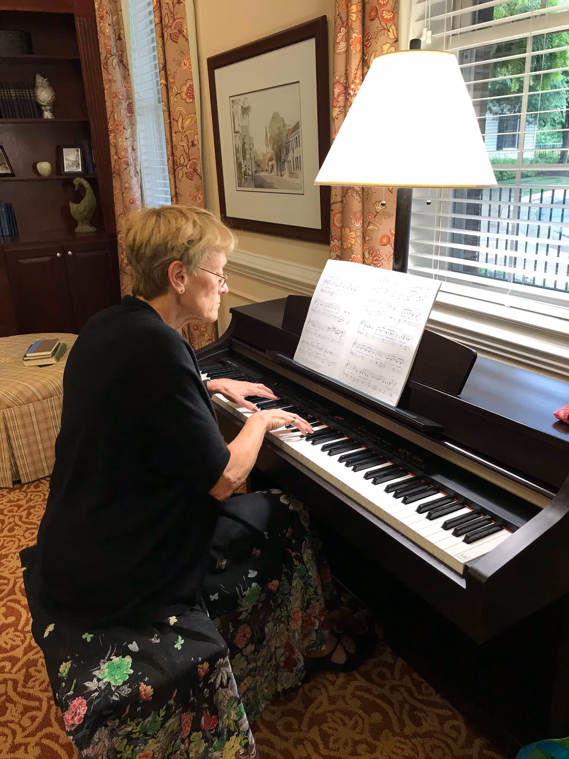 An elderly woman with short blonde hair and glasses is playing a digital piano in a cozy room. The piano has sheet music on the stand and a lamp providing light. Behind her is a window with white blinds and floral curtains, and a framed picture hangs on the wall. A bookshelf and an ottoman with books are visible in the background.