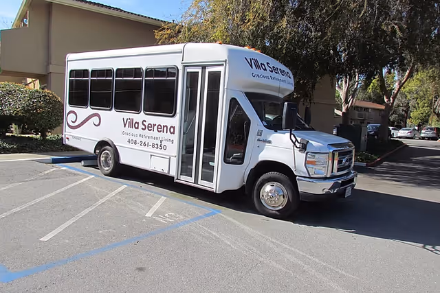 A white Villa Serena retirement community shuttle bus parked in a parking lot by a building and trees.