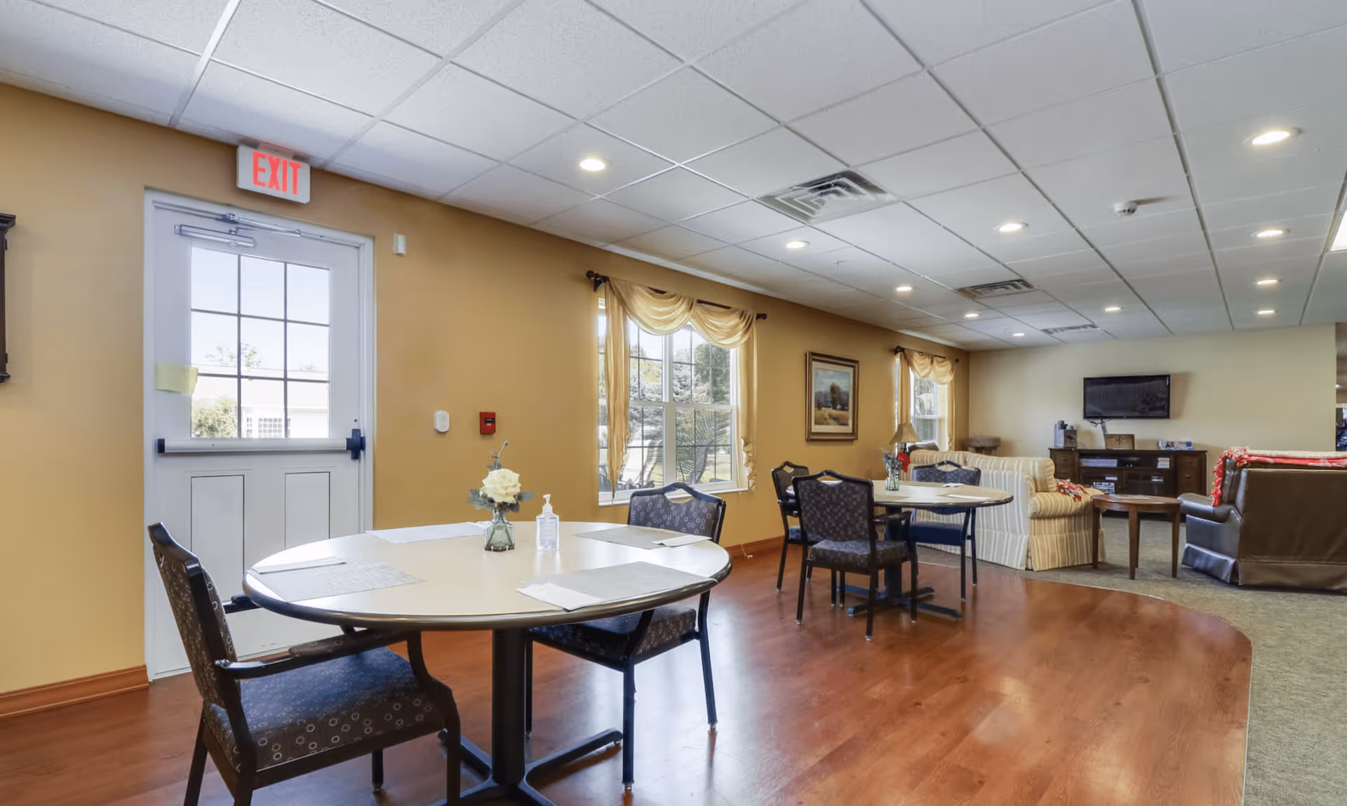 A bright and spacious common area in an assisted living facility featuring round tables with chairs, a vase with flowers, and a bottle of hand sanitizer on one table. The room has large windows with drapes, a television mounted on the wall, comfortable sofas, and warm yellow walls with wood flooring transitioning to carpet.