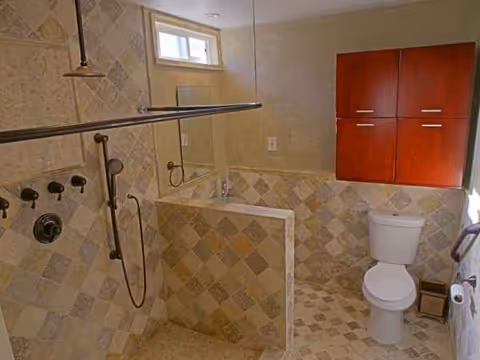 Accessible tiled bathroom with shower, glass partition, toilet, and wooden wall cabinet.