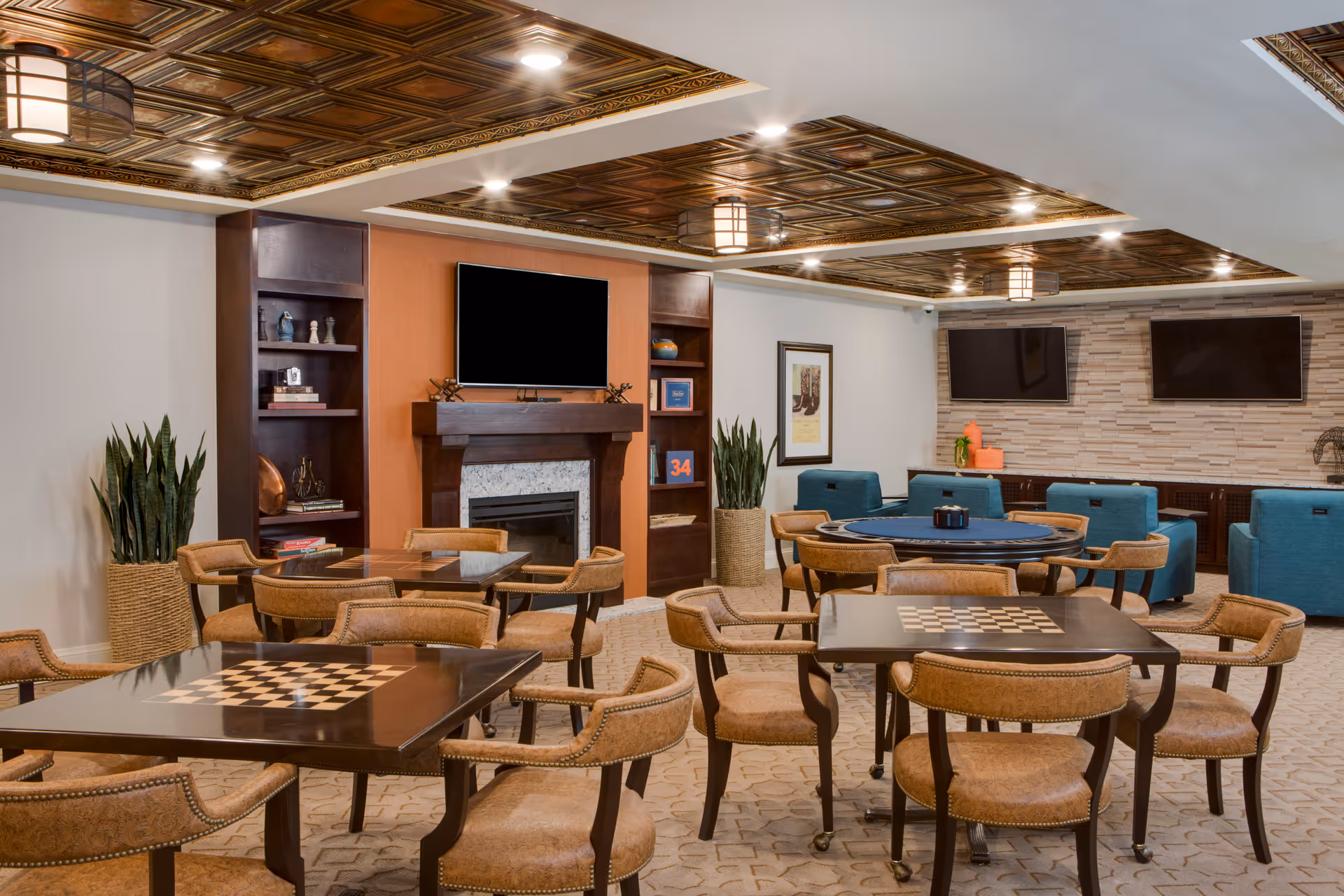 A cozy common area with multiple tables and chairs arranged for socializing and games. The tables have built-in chessboards. The room features a fireplace with a TV mounted above it, built-in shelves with decorative items, two additional wall-mounted TVs, and comfortable blue armchairs. The ceiling has decorative wood panels with recessed lighting, and there are large potted plants adding greenery to the space.