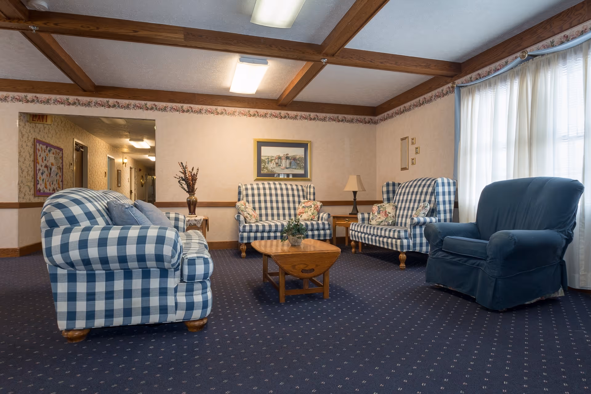 A cozy living room area with blue and white checkered sofas and armchairs arranged around a wooden coffee table. The room has a blue carpet with small patterns, beige walls with floral wallpaper border near the ceiling, wooden ceiling beams, and a large window with sheer white curtains allowing natural light to enter. There is a framed painting on the wall and a table lamp on a side table.