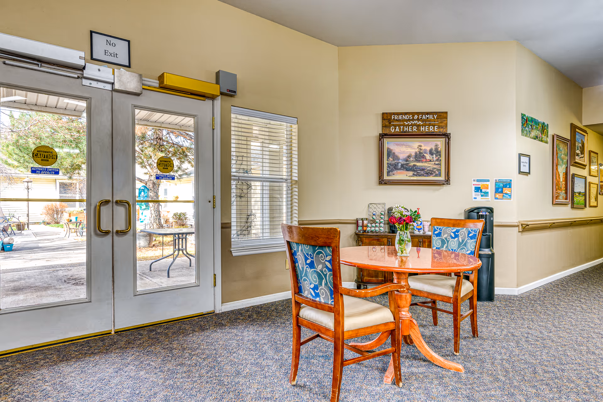 Interior view of a common area in Awbrey Place Assisted Living and Memory Care featuring a round wooden table with two chairs, a vase with flowers on the table, a sideboard with decorative items, and framed artwork on the walls. Double glass doors lead to an outdoor patio area with tables and chairs. A sign above the doors reads 'No Exit' and another sign on the wall says 'Friends & Family Gather Here'.