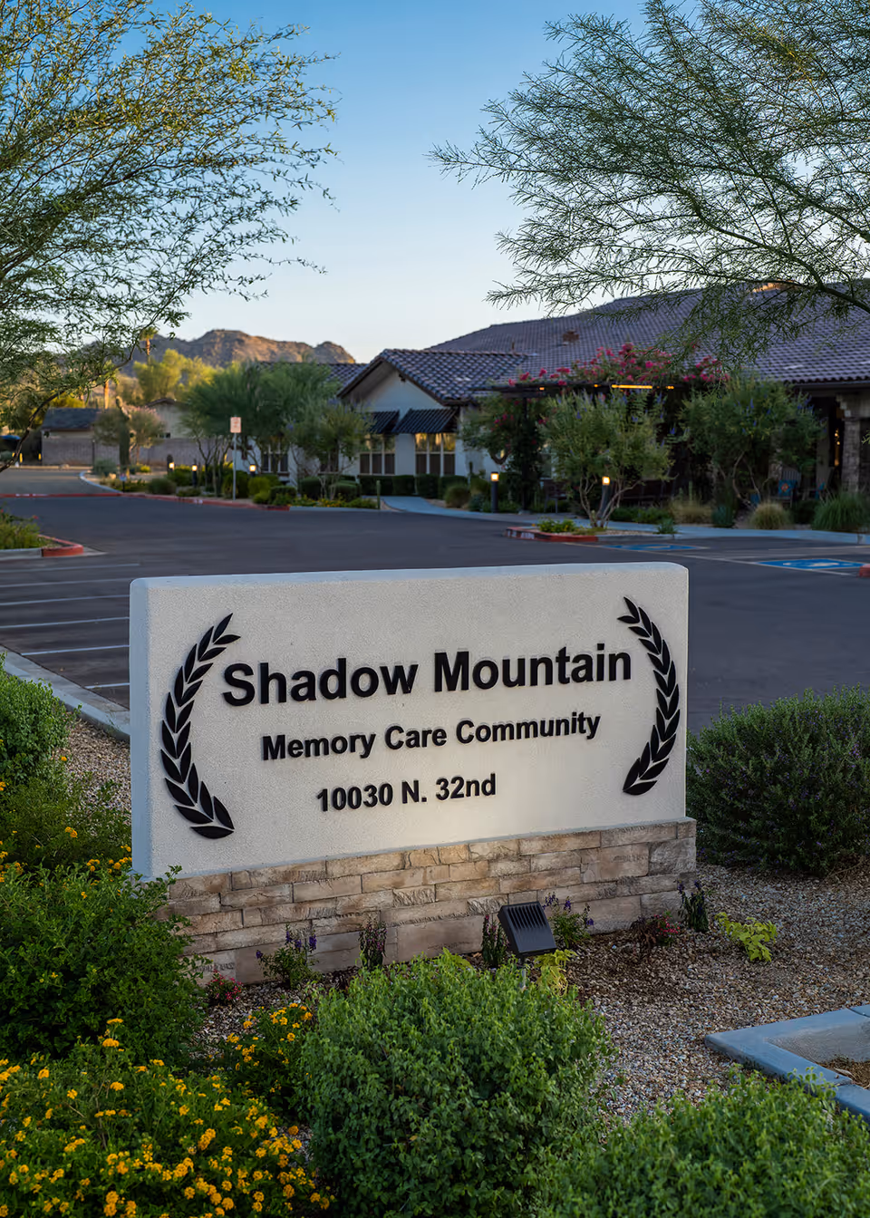 A stone entrance sign reading "Shadow Mountain Memory Care Community 10030 N. 32nd" in front of landscaped shrubs and the facility building.