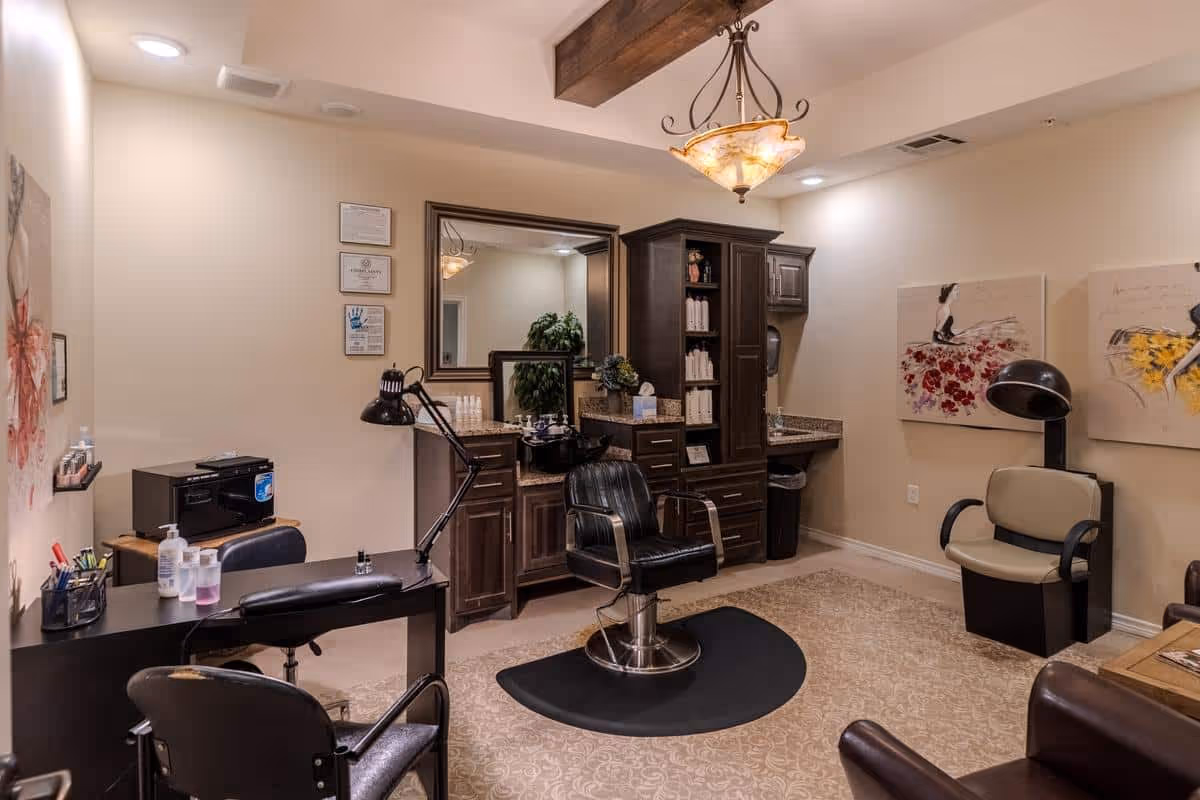 A well-lit salon room with a black salon chair on a protective mat in front of a large mirror and dark wooden cabinetry. The room also contains a desk with manicure supplies, a lamp, and additional seating. The walls are decorated with framed certificates and floral artwork, and the ceiling features a decorative light fixture with wooden beams.