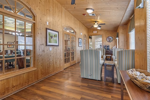 Wood-paneled common lounge with striped armchairs, hardwood floors, ceiling fans, and French doors leading to another room.