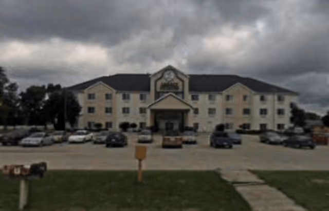 Front exterior view of a three-story beige building with a dark roof, multiple windows, and a central entrance with a covered porch. Several cars are parked in the parking lot in front of the building under a cloudy sky.