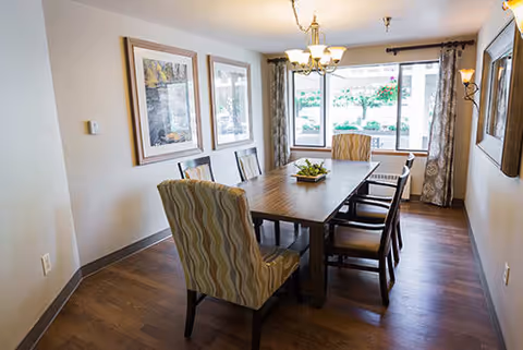 A dining room with a wooden table surrounded by six chairs, two of which have patterned upholstery. The room has wooden flooring, two framed pictures on the wall, a chandelier hanging from the ceiling, and a large window with curtains letting in natural light.