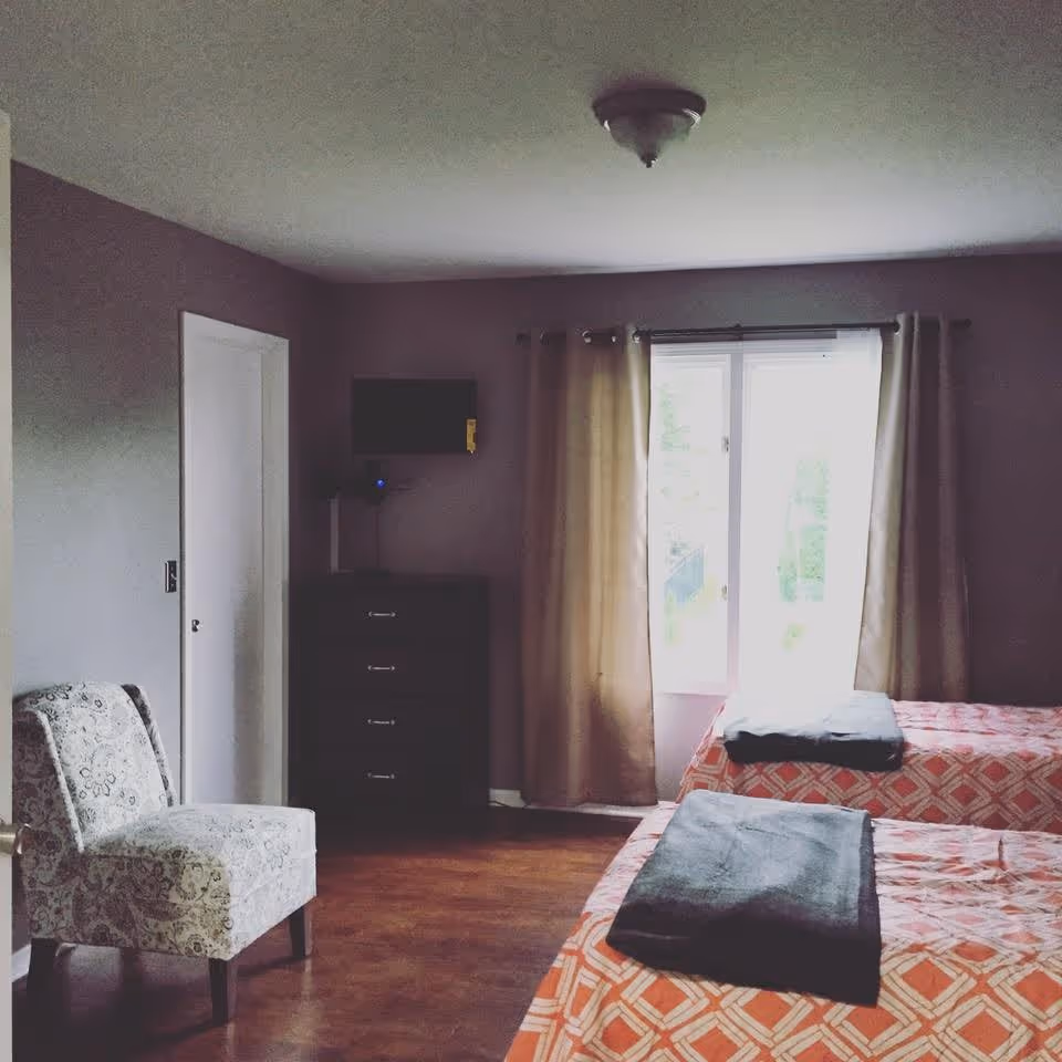 A bedroom with two twin beds covered in orange and white patterned bedspreads, each with a folded dark gray blanket at the foot. There is a window with beige curtains letting in natural light, a patterned armchair to the left, and a dark dresser with a small wall-mounted TV above it against a gray wall.