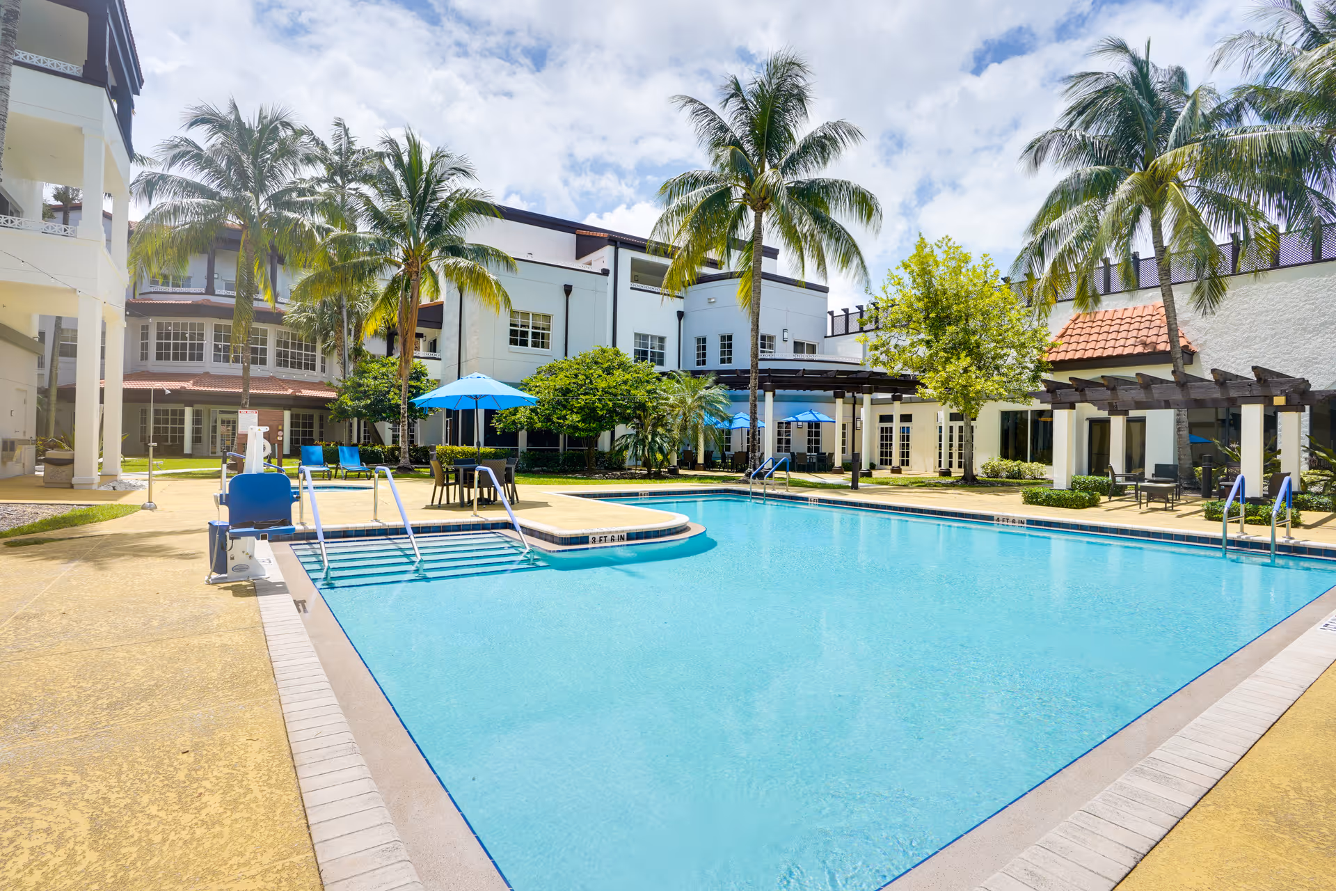 Outdoor swimming pool area at Covenant Living of Florida with clear blue water, poolside chairs, tables with blue umbrellas, palm trees, and surrounding buildings under a partly cloudy sky.