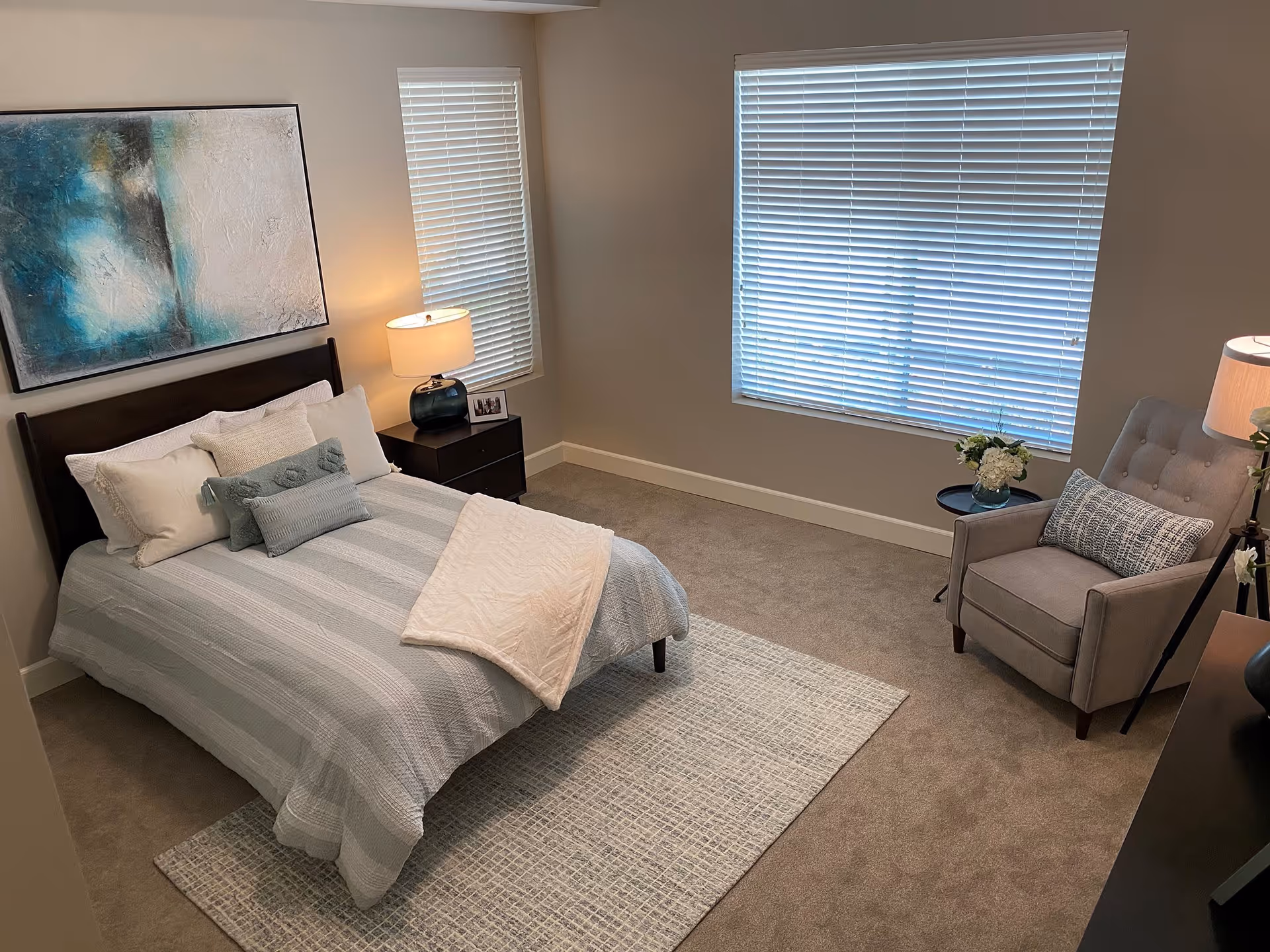 A neatly arranged bedroom with a bed covered in light blue and white striped bedding, multiple pillows, and a white throw blanket. There is a dark wooden nightstand with a lamp and a small framed photo next to the bed. The room has beige carpet, two windows with white blinds, a large abstract painting above the bed, a gray upholstered armchair with a patterned pillow, a small round side table with a vase of flowers, and a floor lamp.