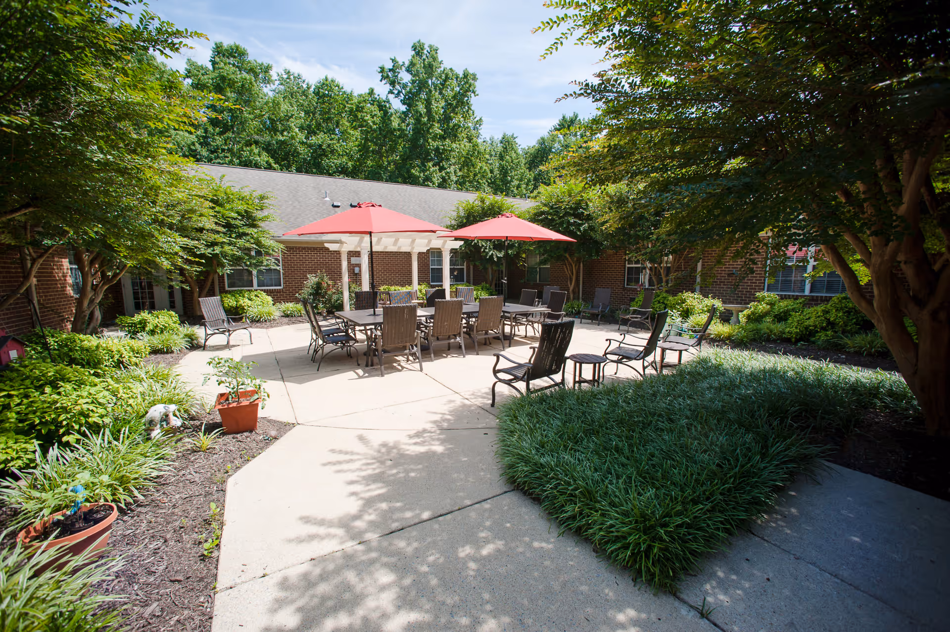Outdoor courtyard area with a concrete patio surrounded by green bushes and trees. There are two large tables with multiple chairs around them, shaded by two red umbrellas. The courtyard is enclosed by a brick building with windows.