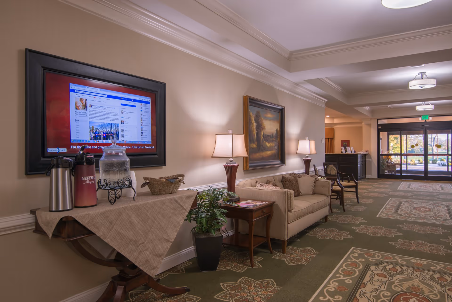A well-lit lobby area with a beige sofa, two table lamps, a side table with magazines, a potted plant, and a table with coffee and water dispensers. A large framed TV screen displays a Facebook page. The floor is carpeted with a patterned design, and glass doors lead outside in the background.