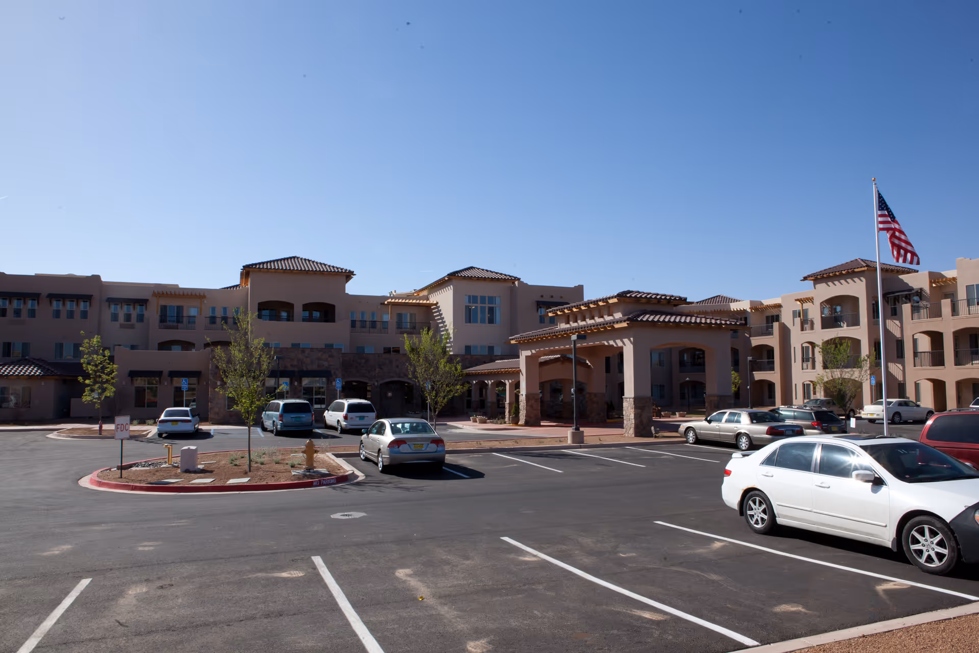 Front entrance and parking lot of a three-story beige retirement living building with an American flag.