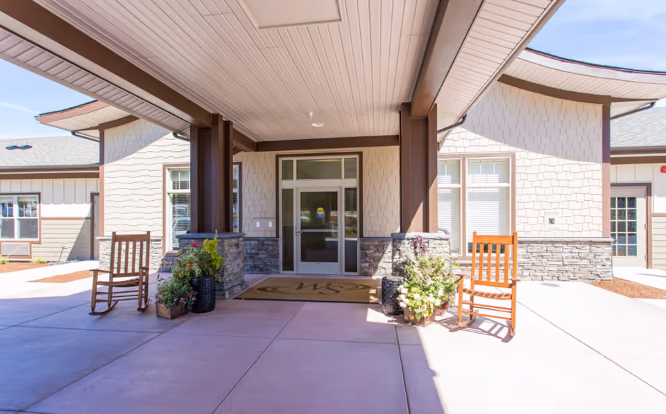 Entrance area of Willamette Springs Memory Care Community featuring a covered porch with two wooden rocking chairs and potted plants on either side of the door. The building exterior has light-colored siding with stone accents and large windows.