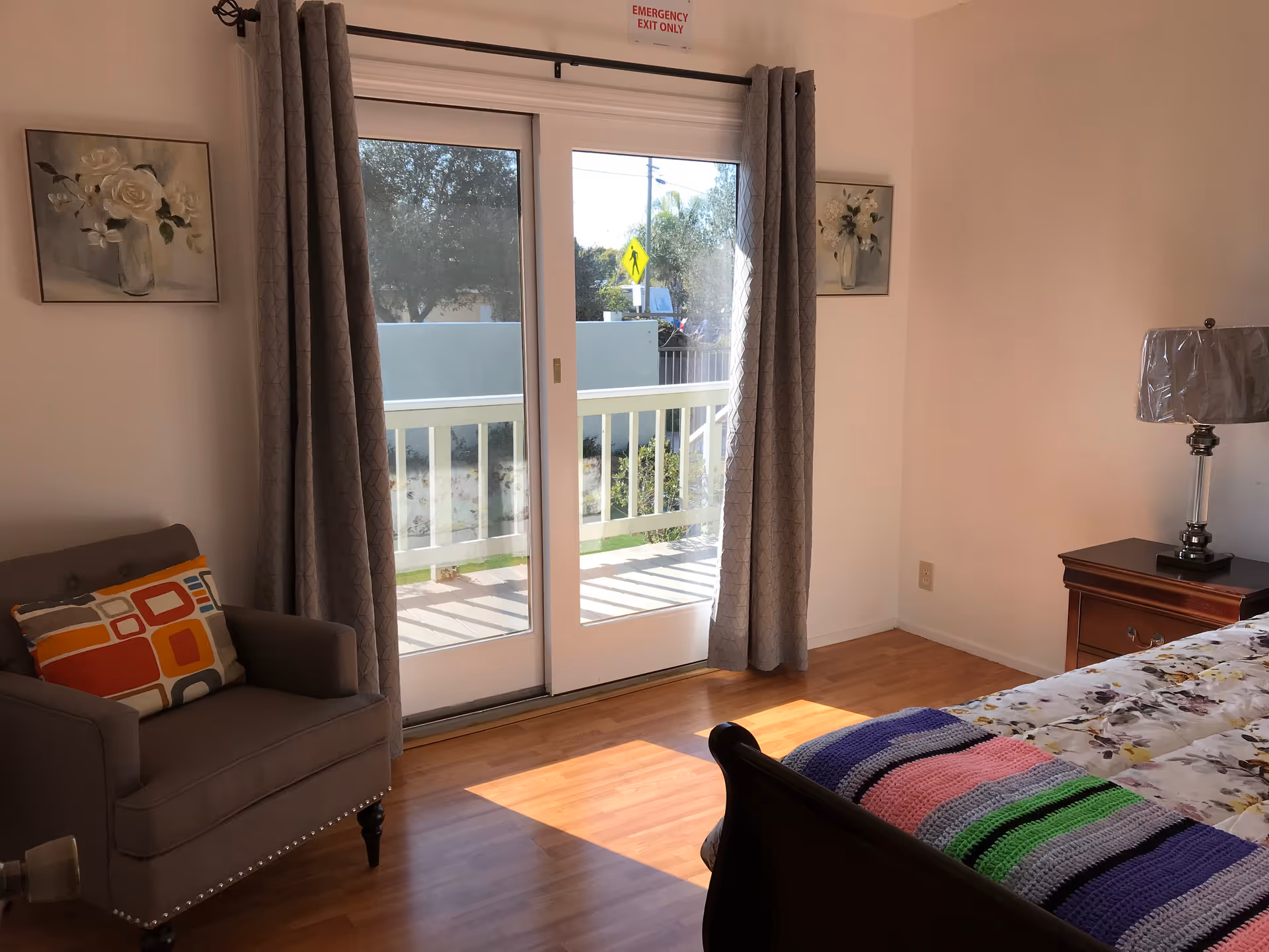 A bright bedroom with wooden flooring featuring a bed with floral bedding and a colorful knitted blanket. There is a wooden nightstand with a lamp on the right side and a gray armchair with a multicolored cushion on the left. Two framed floral paintings hang on the walls beside a sliding glass door with gray curtains, leading to a balcony with a white railing. Outside, trees and a pedestrian crossing sign are visible.