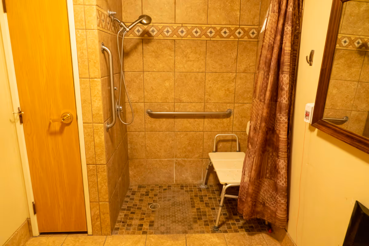 Tiled accessible shower with a handheld showerhead, grab bars, a shower chair and a patterned curtain.