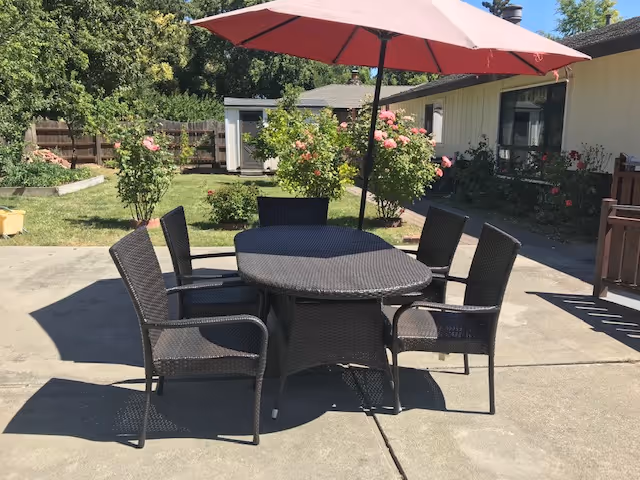 Outdoor patio area with a round wicker table and five matching wicker chairs arranged around it. A large red umbrella provides shade over the table. The patio is surrounded by green grass, flowering bushes, and a wooden fence. A single-story building with windows and flower beds is visible in the background.
