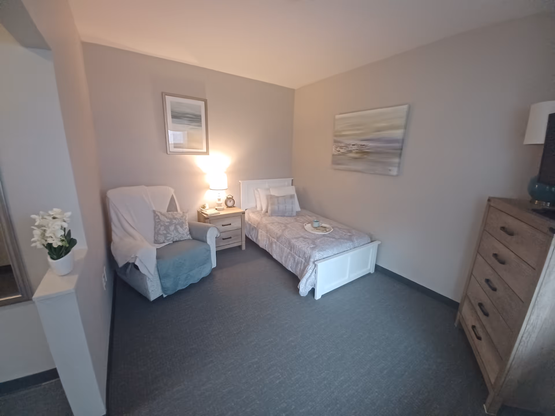 A small, cozy bedroom with a single bed covered in a light gray patterned comforter and pillows. Next to the bed is a wooden nightstand with a lamp, an alarm clock, and a small decorative item. To the left of the nightstand is a comfortable armchair draped with a white throw and a decorative pillow. On the right side of the room is a wooden dresser with a lamp and a TV on top. The walls are painted light gray and decorated with two framed paintings. A small white flower pot with white flowers is placed on a ledge near the armchair.
