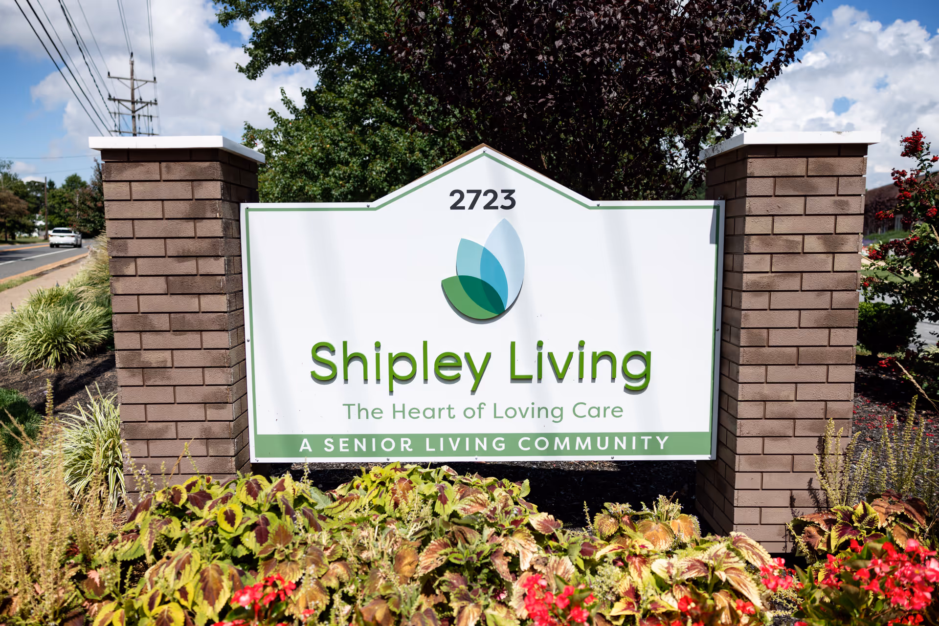 Outdoor sign for Shipley Living, a senior living community, with the address number 2723. The sign is white with green text and a leaf logo, surrounded by plants and flowers, with a clear sky and trees in the background.
