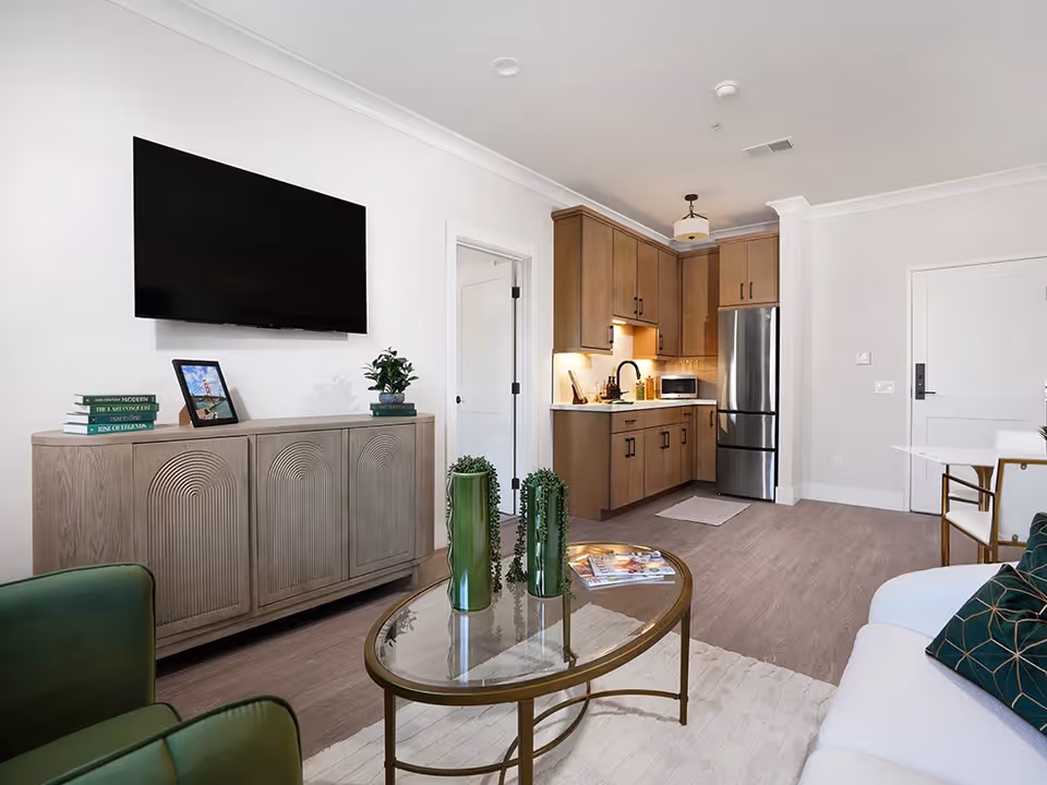 A modern living room and kitchen area in a senior living facility. The living room features a wall-mounted flat-screen TV above a wooden cabinet with decorative items and books. There is a glass coffee table with green vases and a white rug on the floor. The kitchen has wooden cabinets, a stainless steel refrigerator, a microwave, and a sink with under-cabinet lighting. The space has light-colored walls and wood flooring.