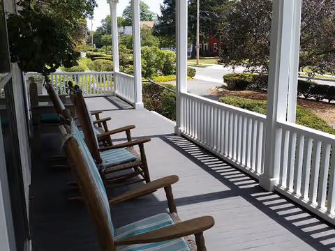 Front porch with several wooden rocking chairs and white railing overlooking a landscaped street.